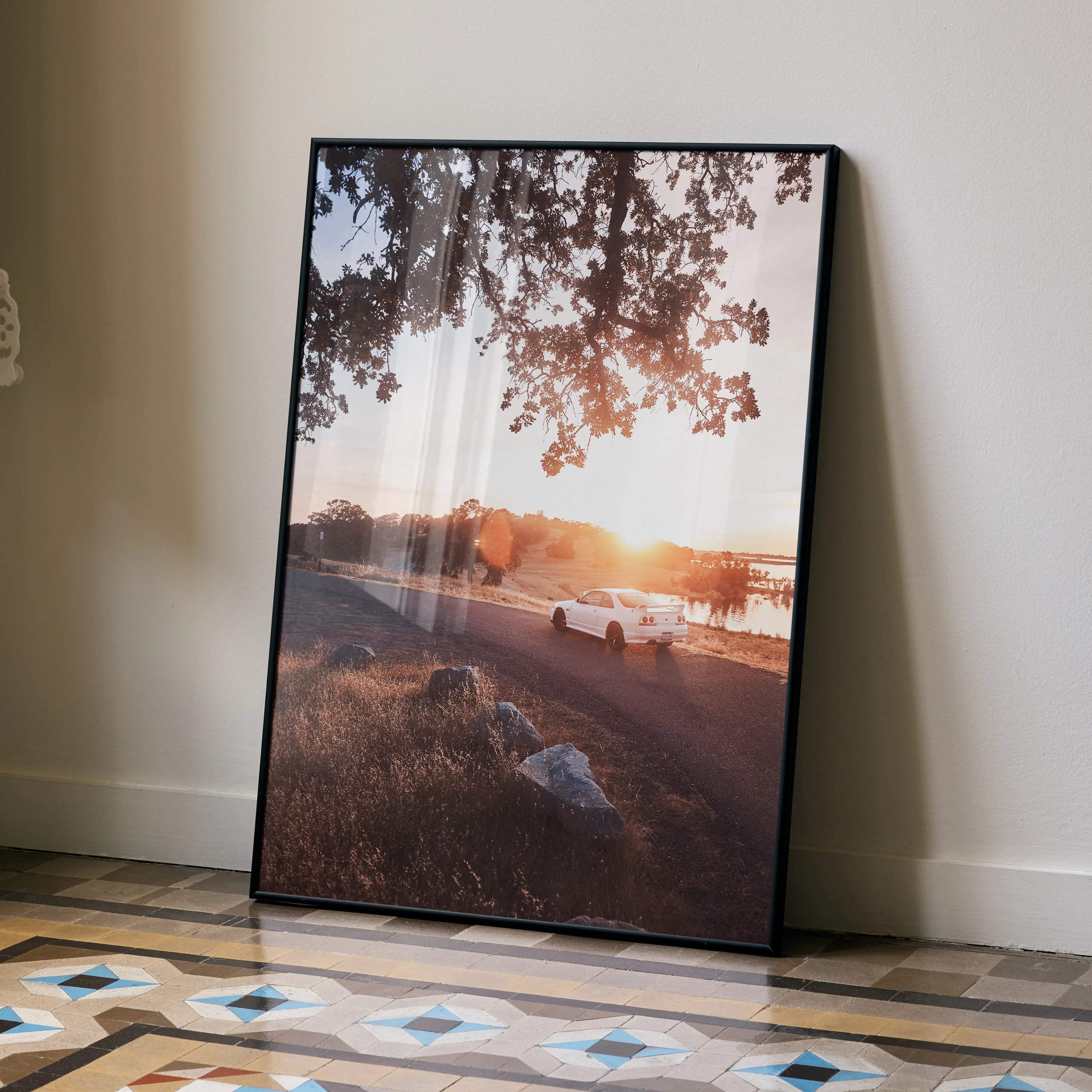 Black-framed photographic print of a white Nissan Skyline R33 GTS-T driving on a sunlit road under tree branches