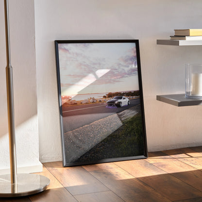 Nissan 370z driving on racetrack at sunset poster wall art in black frame on floor.