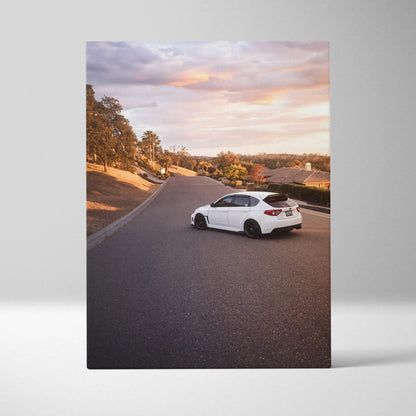 Vertical canvas wall art featuring a white Subaru WRX STI on a scenic road during sunset.