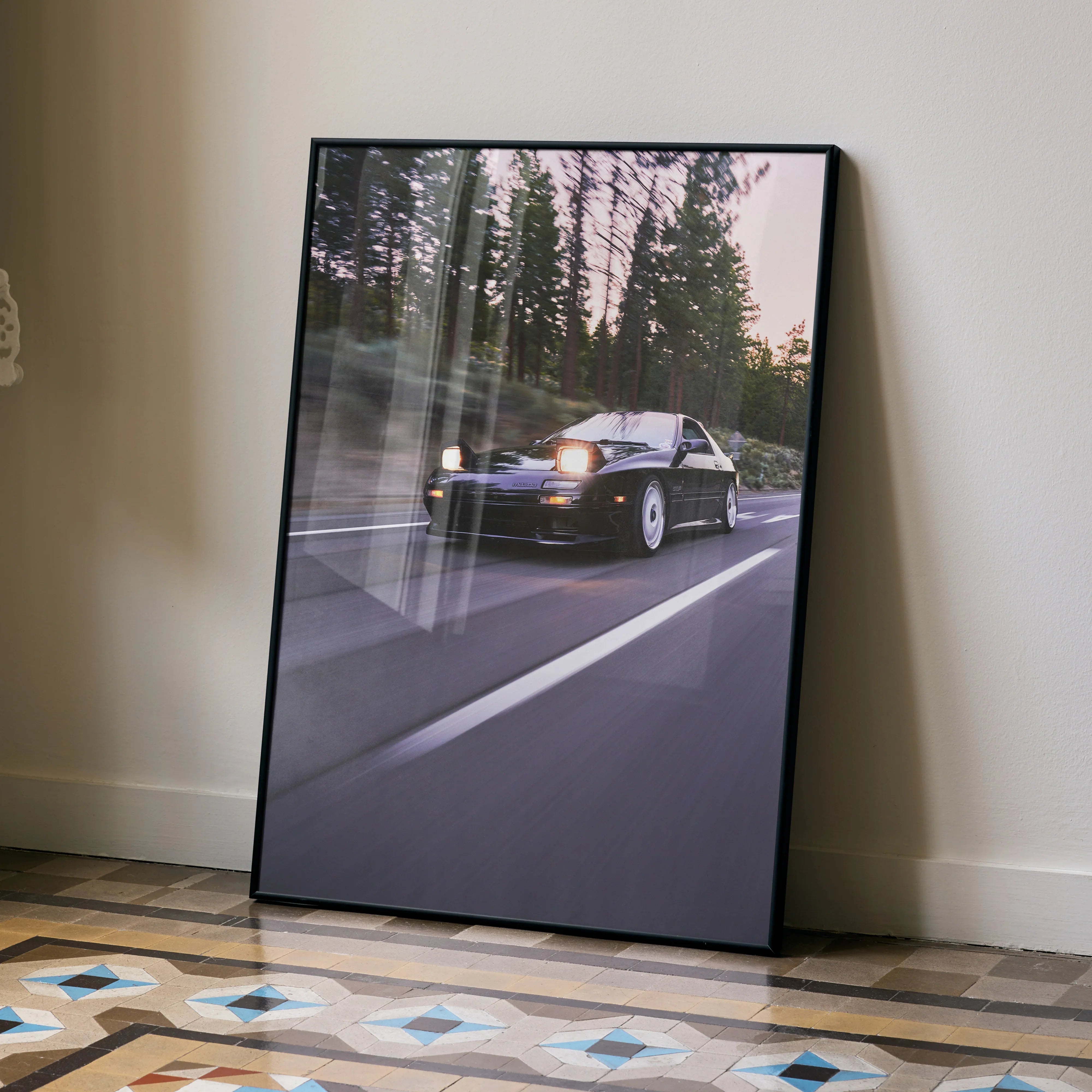 Mazda FC RX7 driving on forest road poster in black frame wall art leaning on floor.