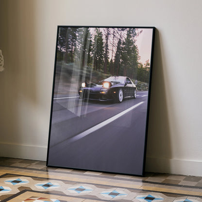 Mazda FC RX7 driving on forest road poster in black frame wall art leaning on floor.