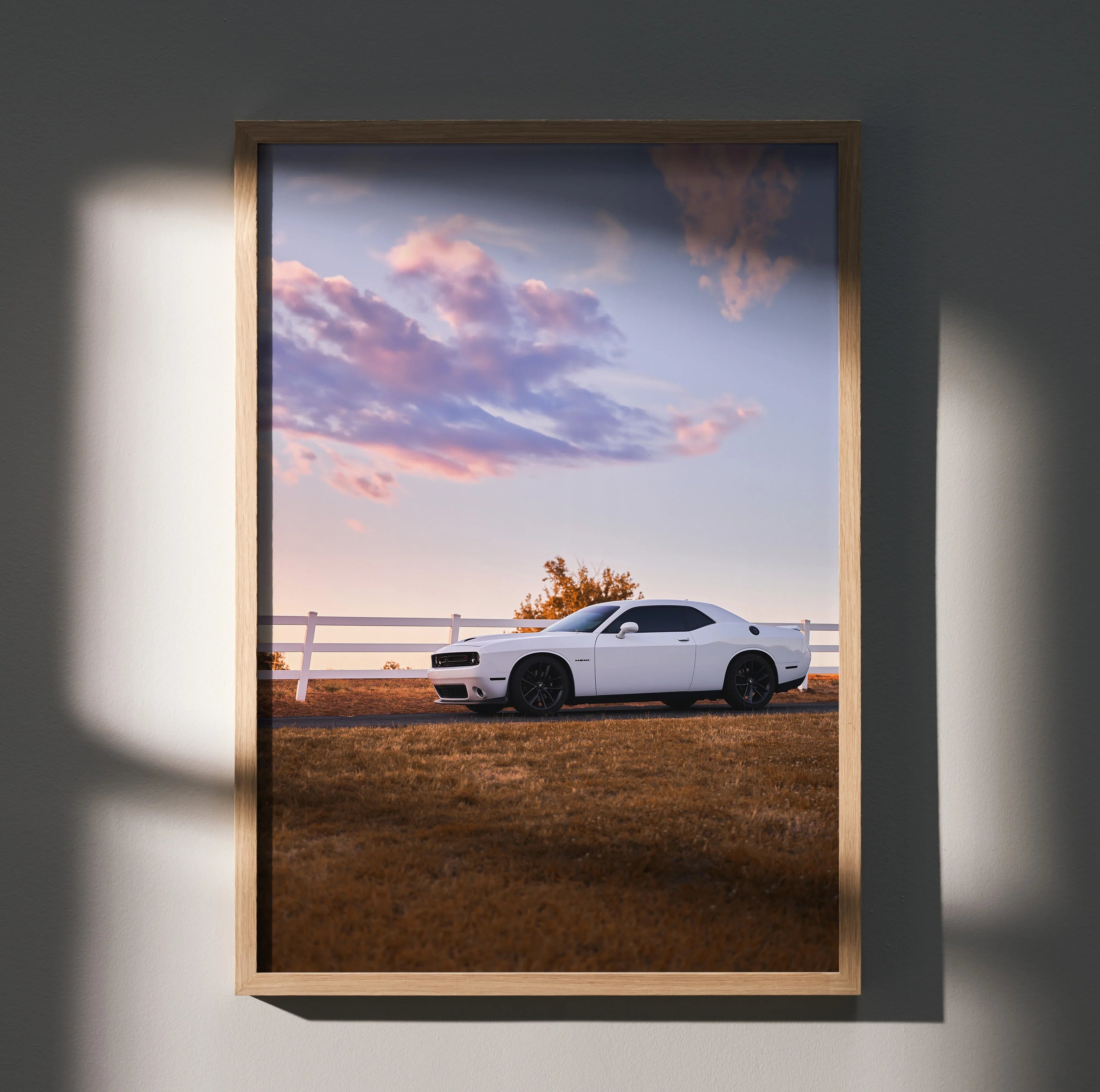 Dodge Challenger parked by white fence under colorful sunset sky poster wall art in wooden frame.
