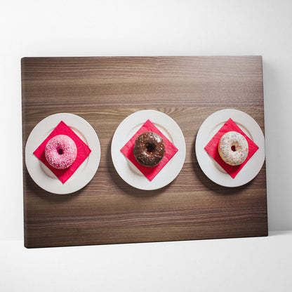 Canvas wall art featuring three glazed donuts—pink, chocolate, and white with sprinkles—each on white plates with red.