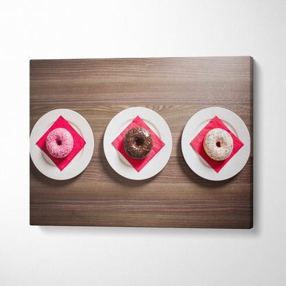 Canvas wall art featuring three vibrant glazed donuts on white plates with red napkins on wood table decor.