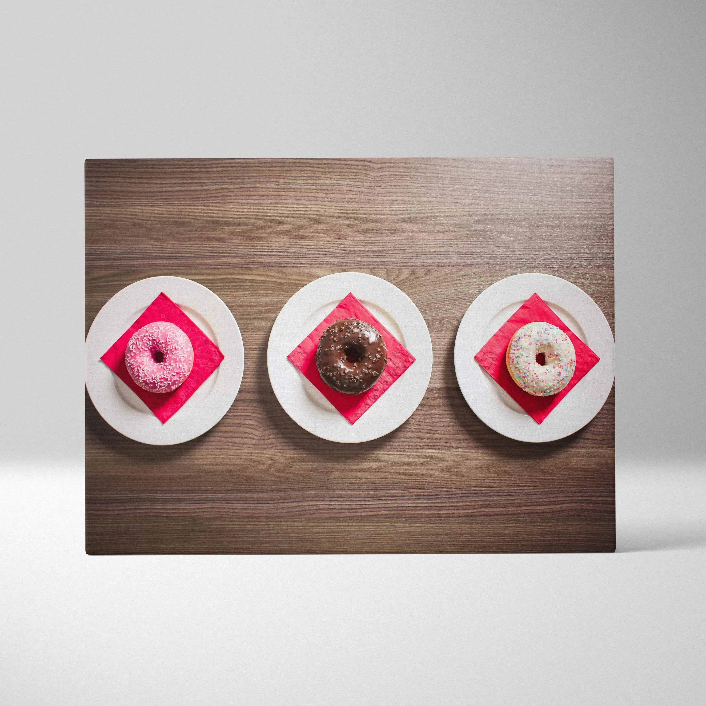 Canvas art print featuring three vibrant glazed donuts on white plates with red napkins, styled on a wooden surface.
