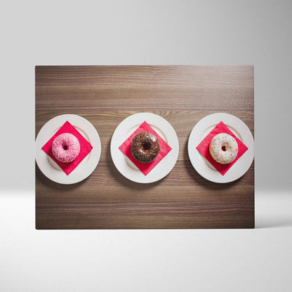 Canvas art print featuring three vibrant glazed donuts on white plates with red napkins, styled on a wooden surface.
