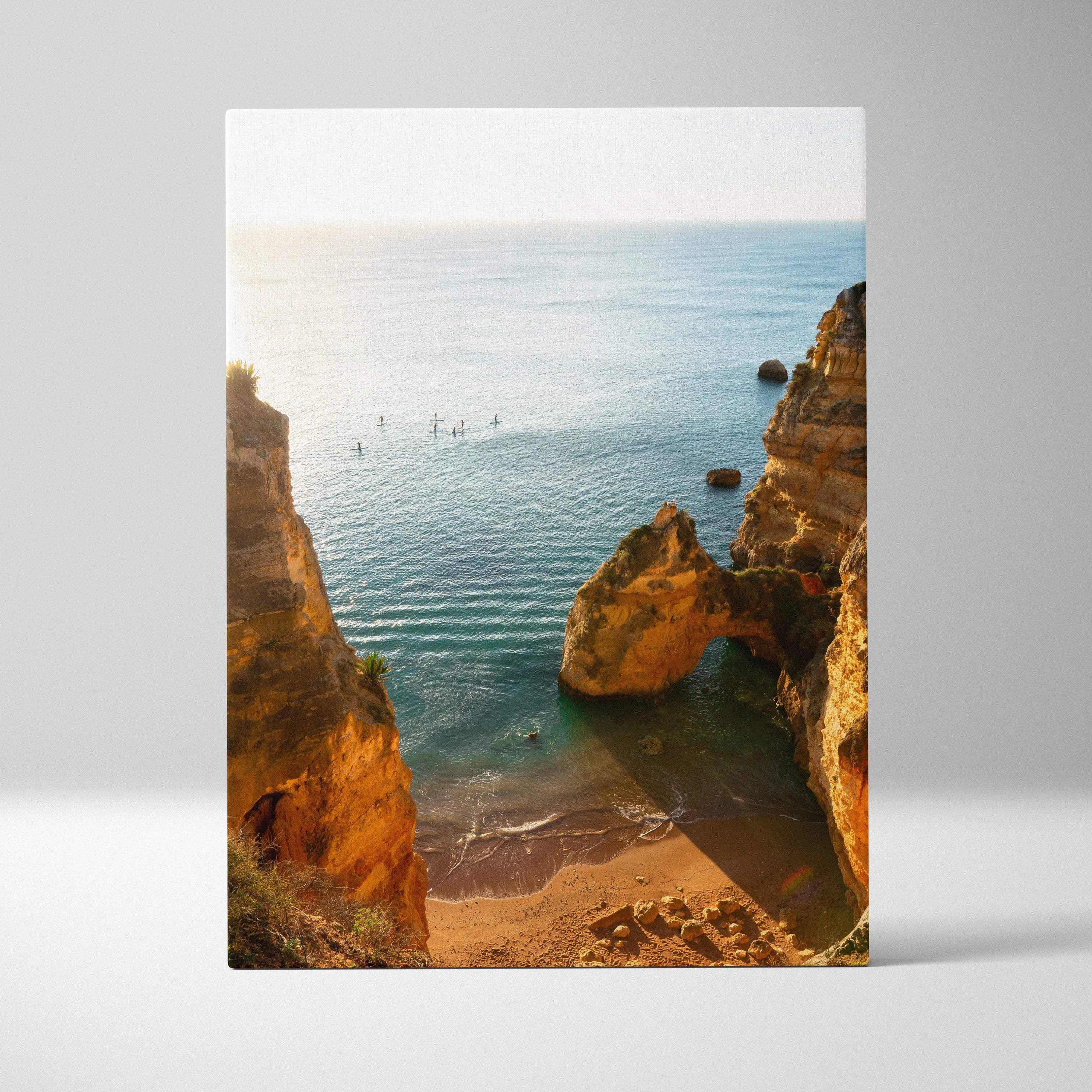 Canvas wall art of a sunlit beach scene with rocky cliffs and calm ocean water.