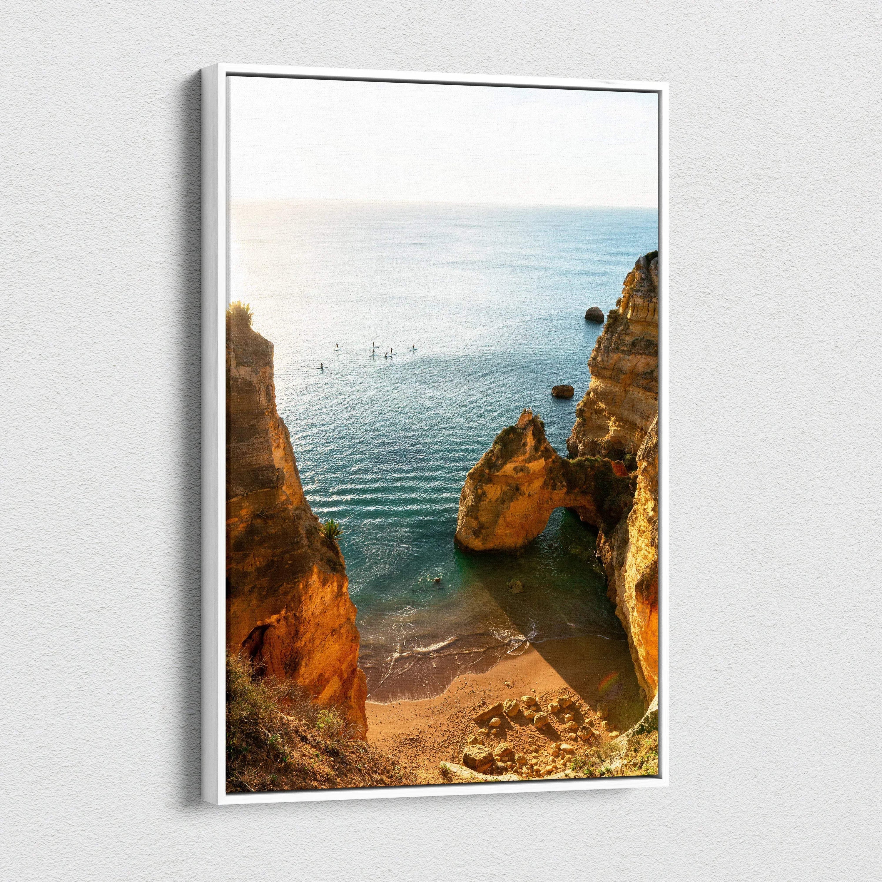 Framed canvas wall art featuring a sunlit beach scene with rocky cliffs and calm blue water.