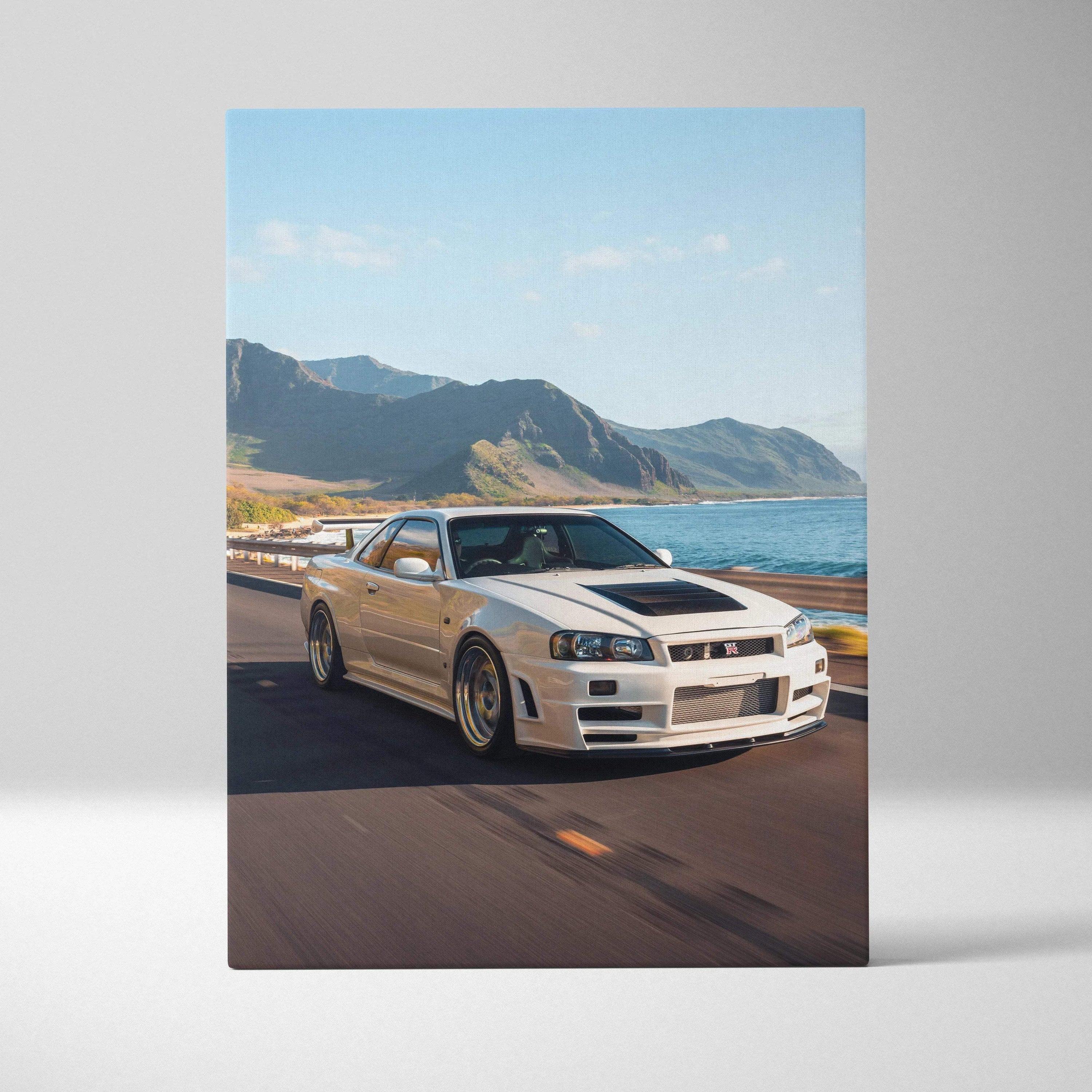Vertical canvas art featuring a white Nissan R34 GTR Skyline driving along a coastal road with mountains and ocean backdrop.