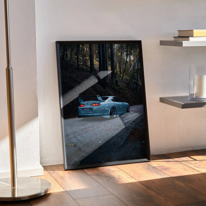 Toyota Supra Mk4 poster wall art with rear view of blue car on forest road, framed and leaning on floor.