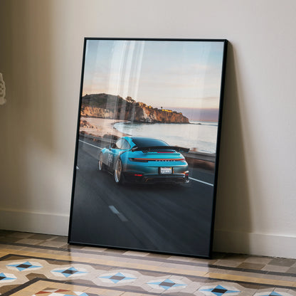 Porsche 911 driving on coastal road at sunset poster wall art with black frame.
