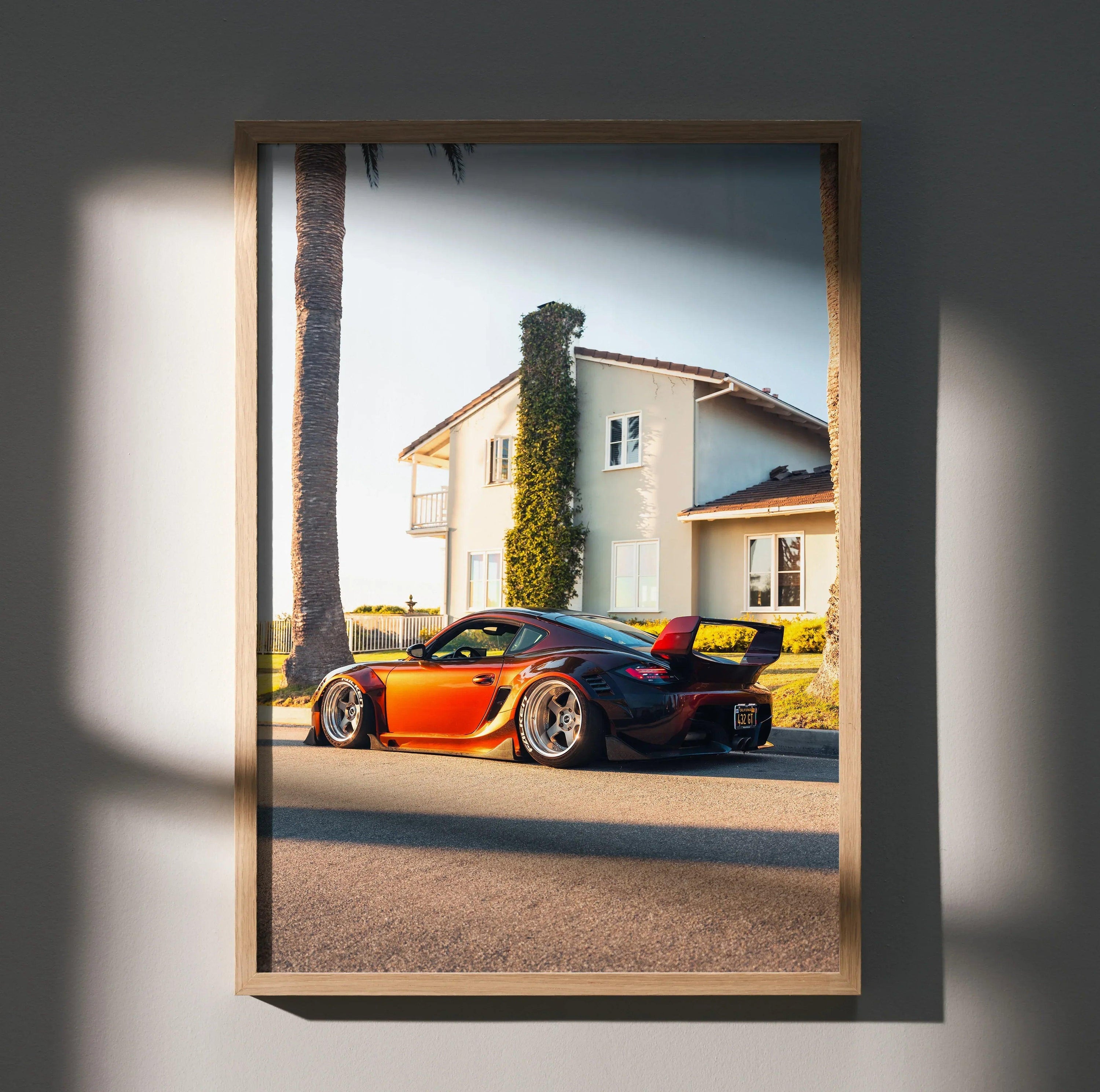 Porsche Cayman poster showing an orange and black sports car parked by a house wall art in wooden frame.