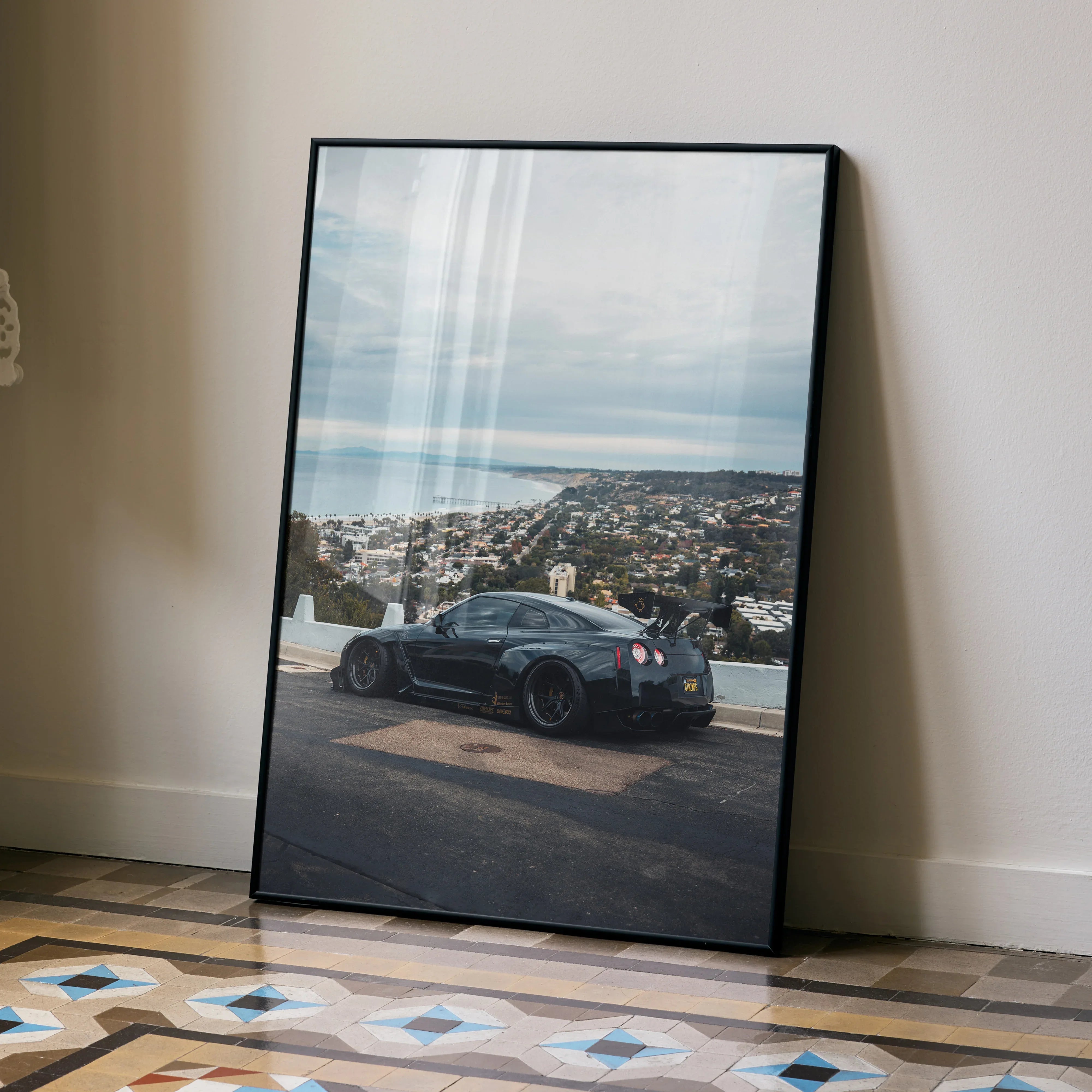 Nissan GTR R35 poster wall art featuring black sports car overlooking coastal cityscape under cloudy sky.