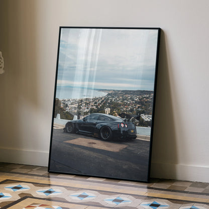 Nissan GTR R35 poster wall art featuring black sports car overlooking coastal cityscape under cloudy sky.