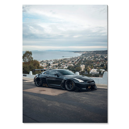 Black Nissan R35 GTR on coastal road with cityscape background wall art poster.