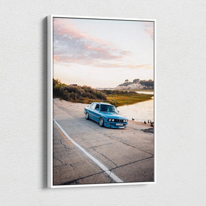 Vertical canvas wall art featuring a blue BMW E28 5 Series parked by water at sunset.