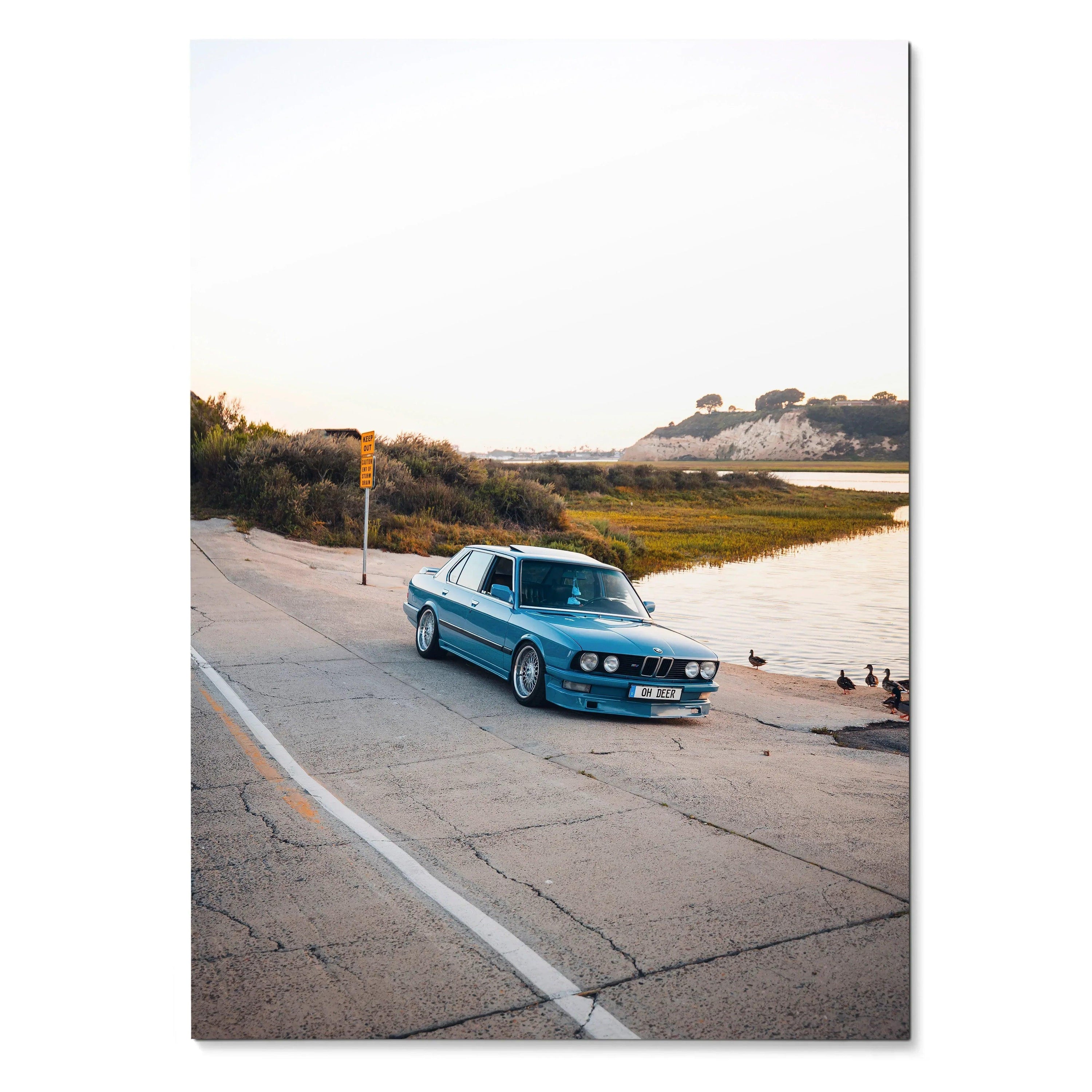 Blue BMW E28 classic car parked by a waterfront road at sunset poster wall art.