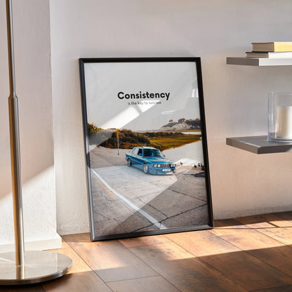 Motivational wall art poster featuring blue BMW E30 on scenic road with "Consistency" text, black frame.