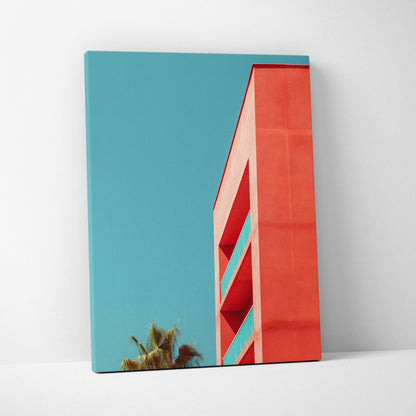 Vertical canvas wall art featuring modern red building against clear blue sky and palm trees in background.