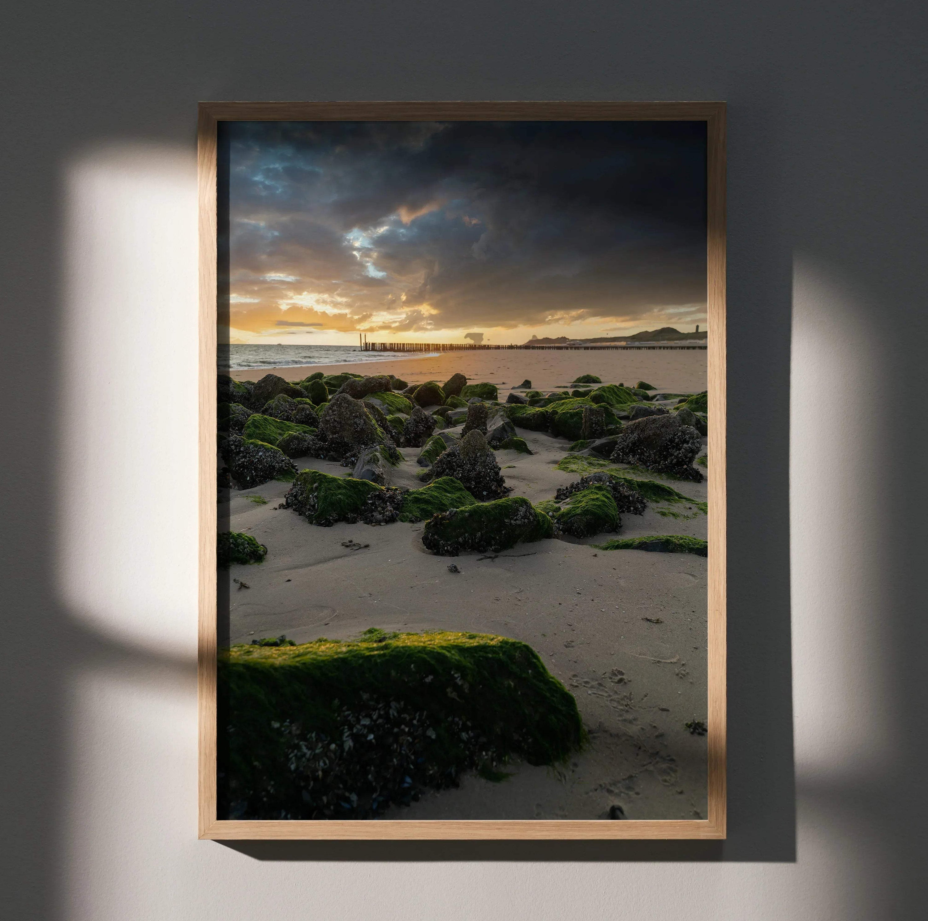 Vertical poster wall art featuring a serene ocean sunset with moss-covered rocks on sandy shore.
