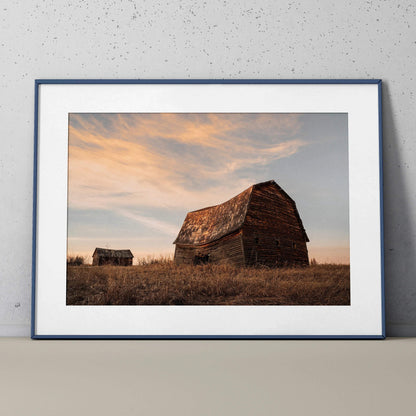 Rustic barn and small building at sunset with soft clouds, framed wall art poster in blue frame.