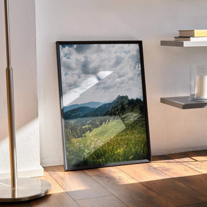 Majestic mountain landscape poster with green hills and cloudy sky in black frame leaning on floor wall art.