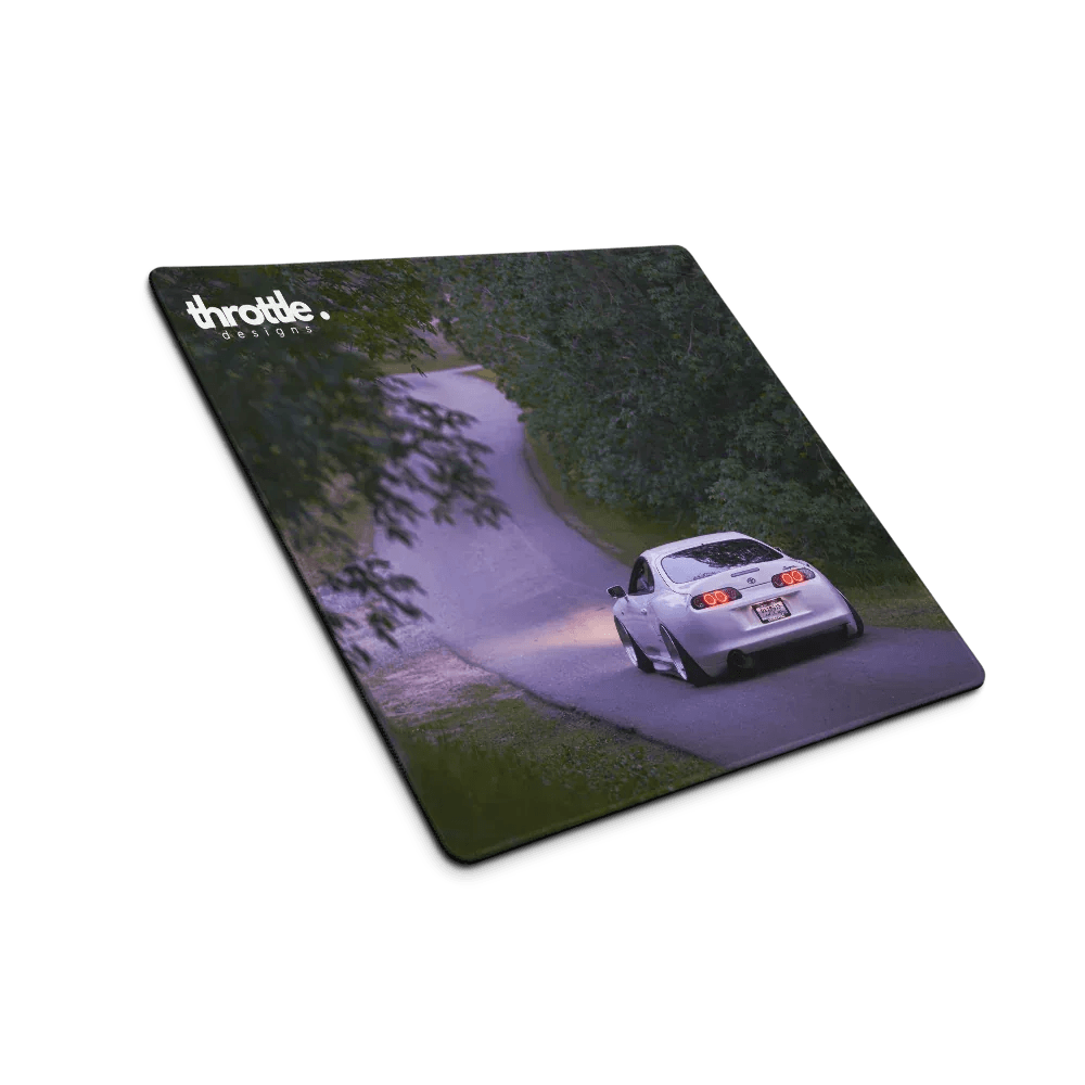 Gaming mouse pad featuring a rear view of a white Toyota Supra Mk4 driving on a winding tree-lined road at dusk.