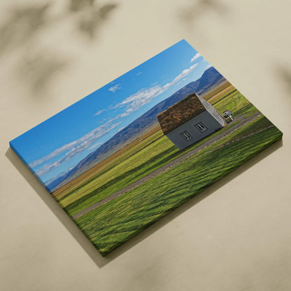 Canvas wall art featuring a rustic building in green fields with mountains under a blue sky.