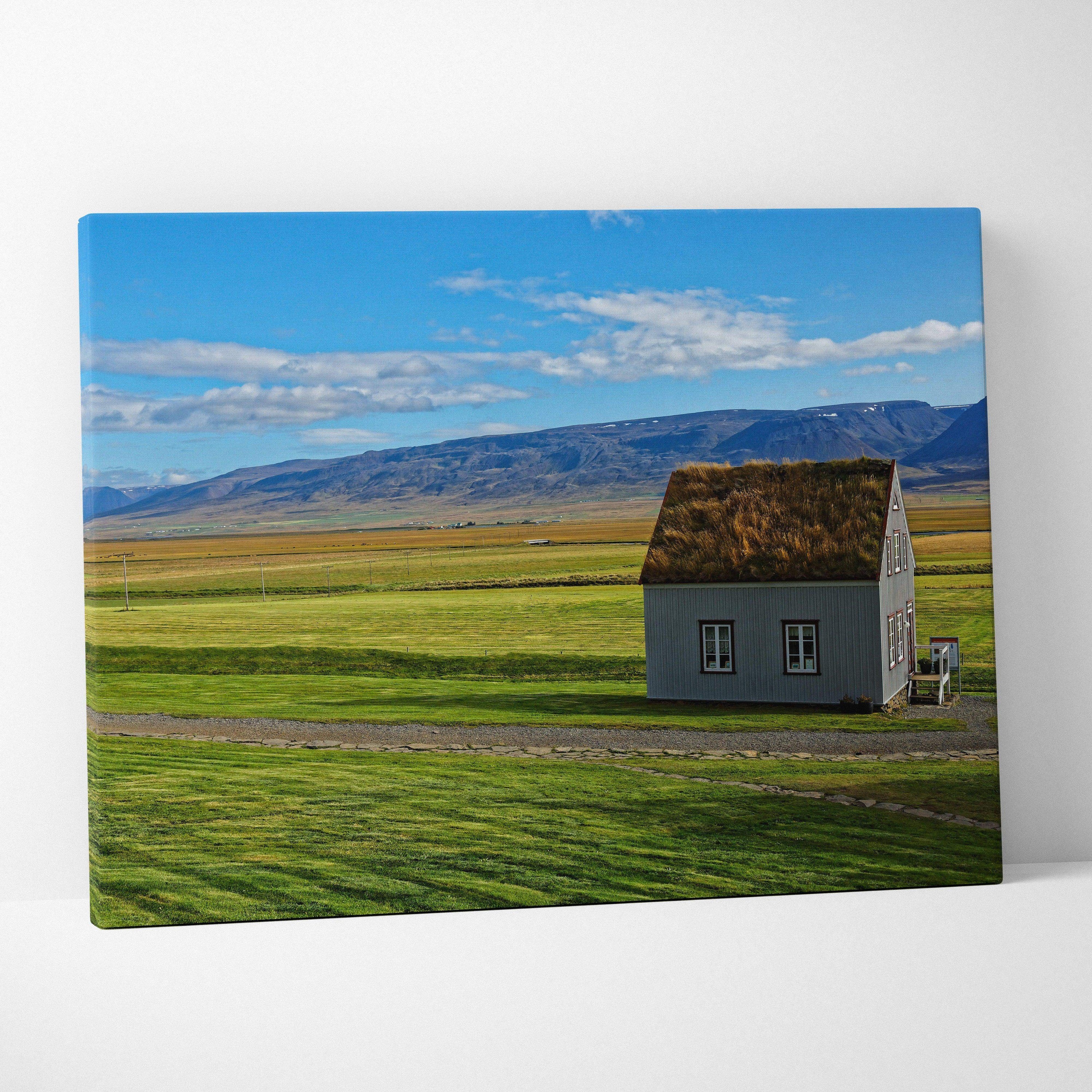 Canvas wall art featuring a small rustic house in green fields under a blue sky with mountains in the background.
