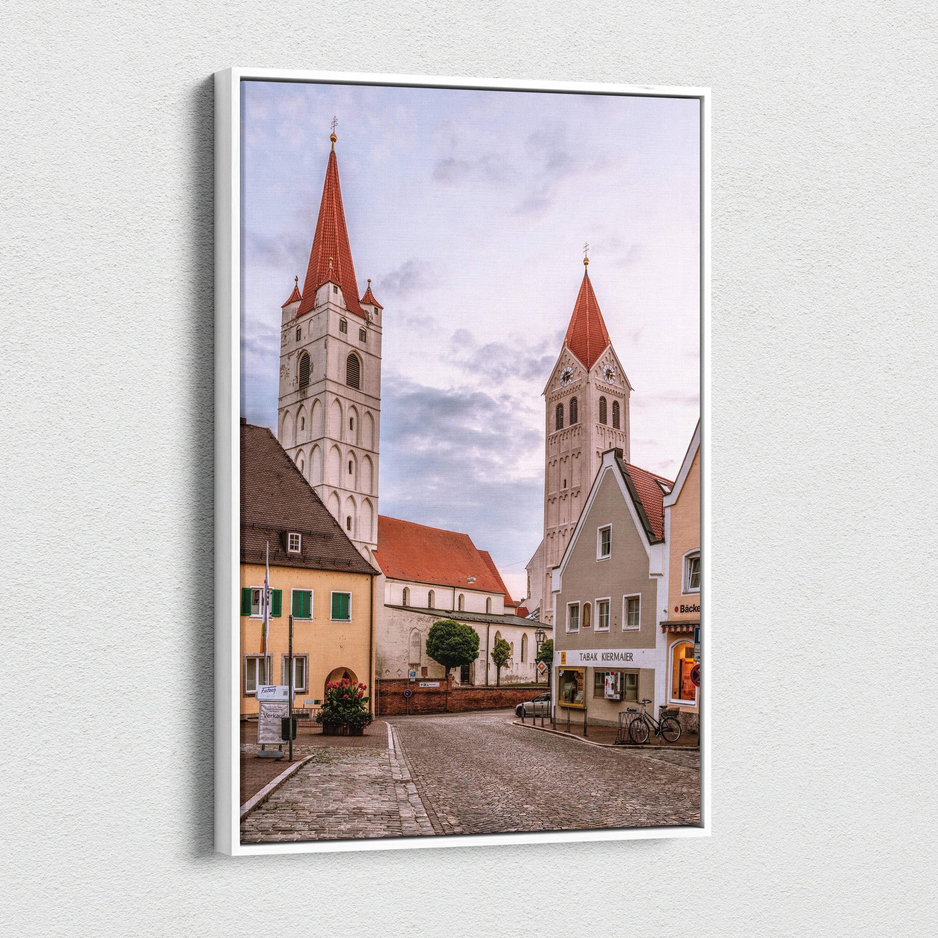 Vertical canvas wall art featuring quaint European village buildings with two tall church steeples under a cloudy sky.