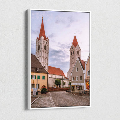 Vertical canvas wall art featuring quaint European village buildings with two tall church steeples under a cloudy sky.