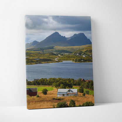 Vertical canvas wall art of majestic mountains over a lake with houses in a grassy field beneath cloudy sky.