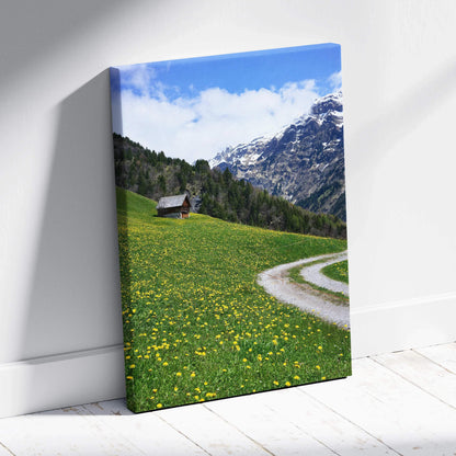 Mountain landscape canvas wall art with green meadow, wildflowers, cabin, and snowy peaks under blue sky.