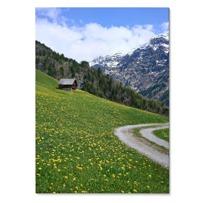 Vertical poster wall art featuring a green hillside with yellow flowers, a small cabin, and snow-capped mountains under a.