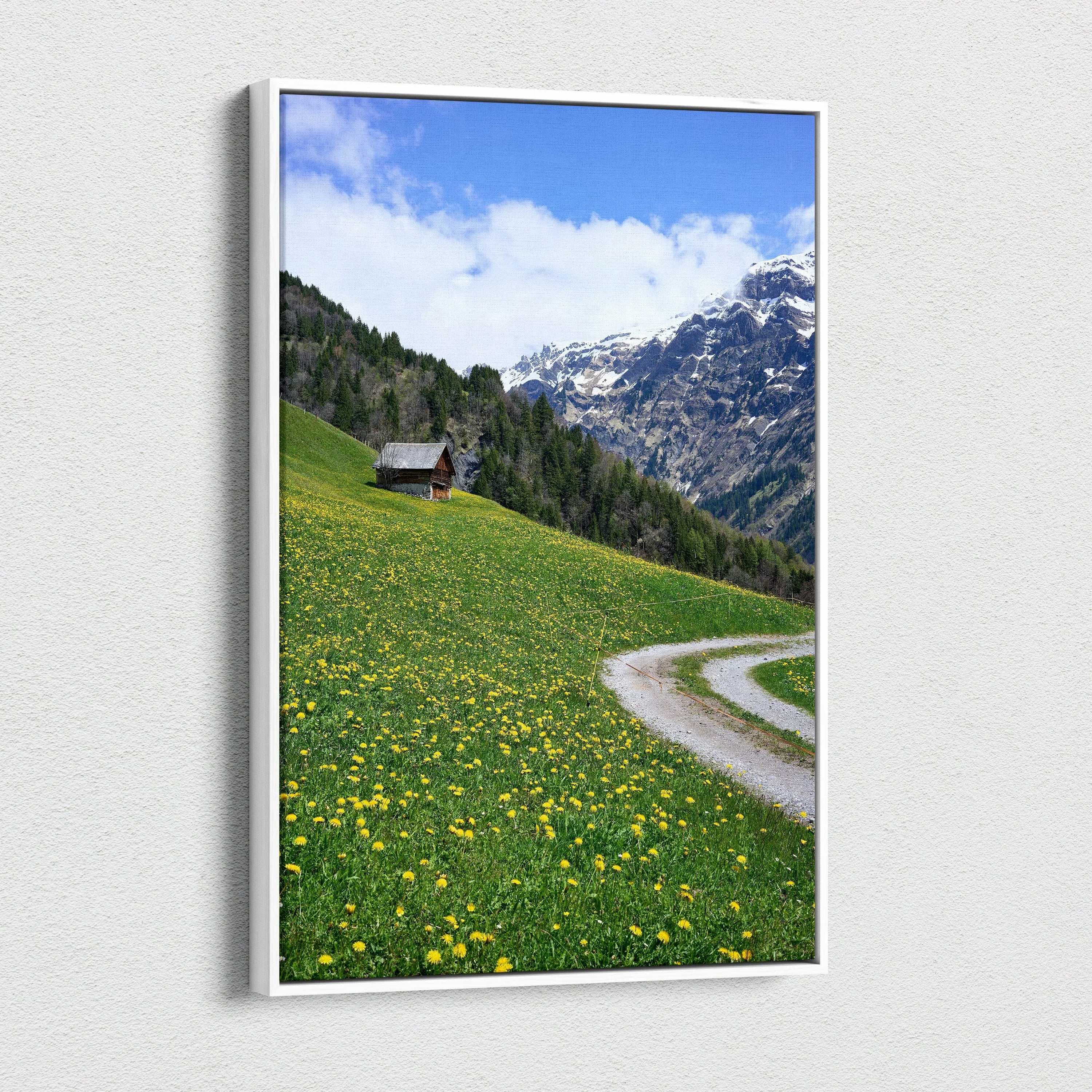 Canvas wall art featuring a green meadow with yellow flowers, winding path, and snow-capped mountain background.