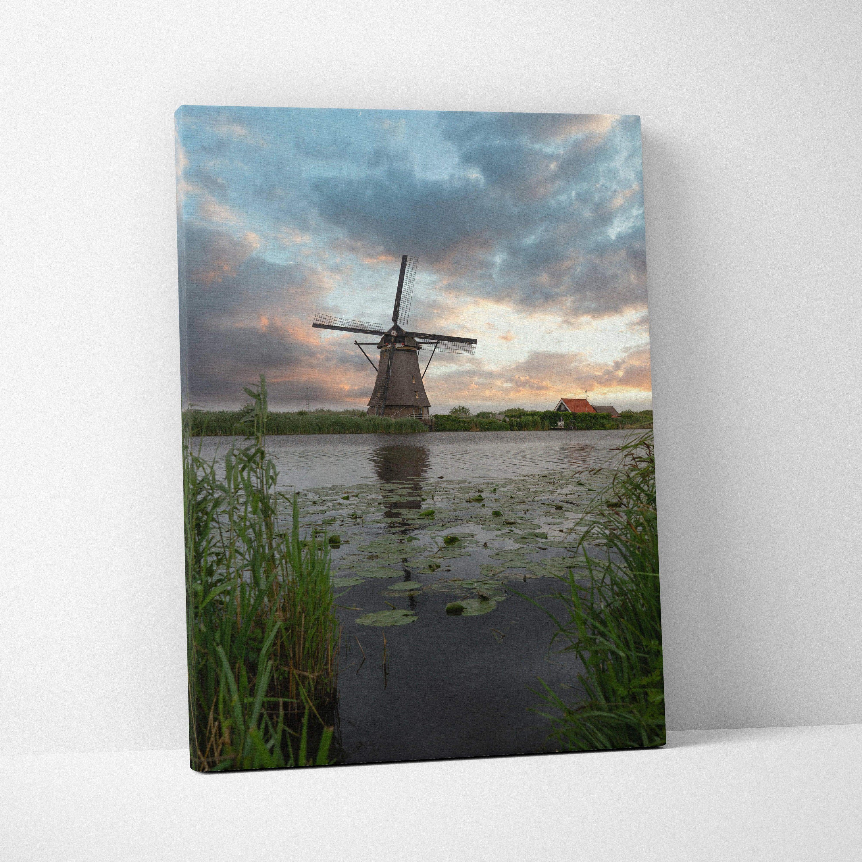 Vertical canvas wall art featuring a windmill silhouette at sunset over a reflective pond with greenery.