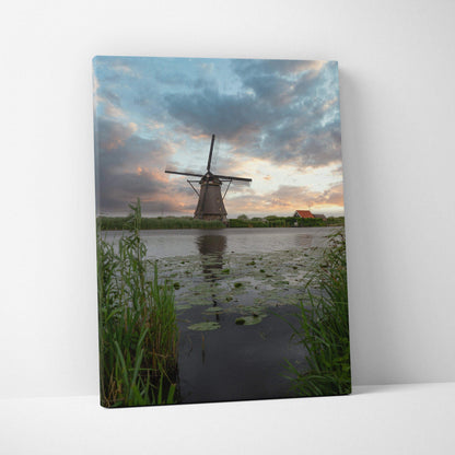 Vertical canvas wall art featuring a windmill silhouette at sunset over a reflective pond with greenery.