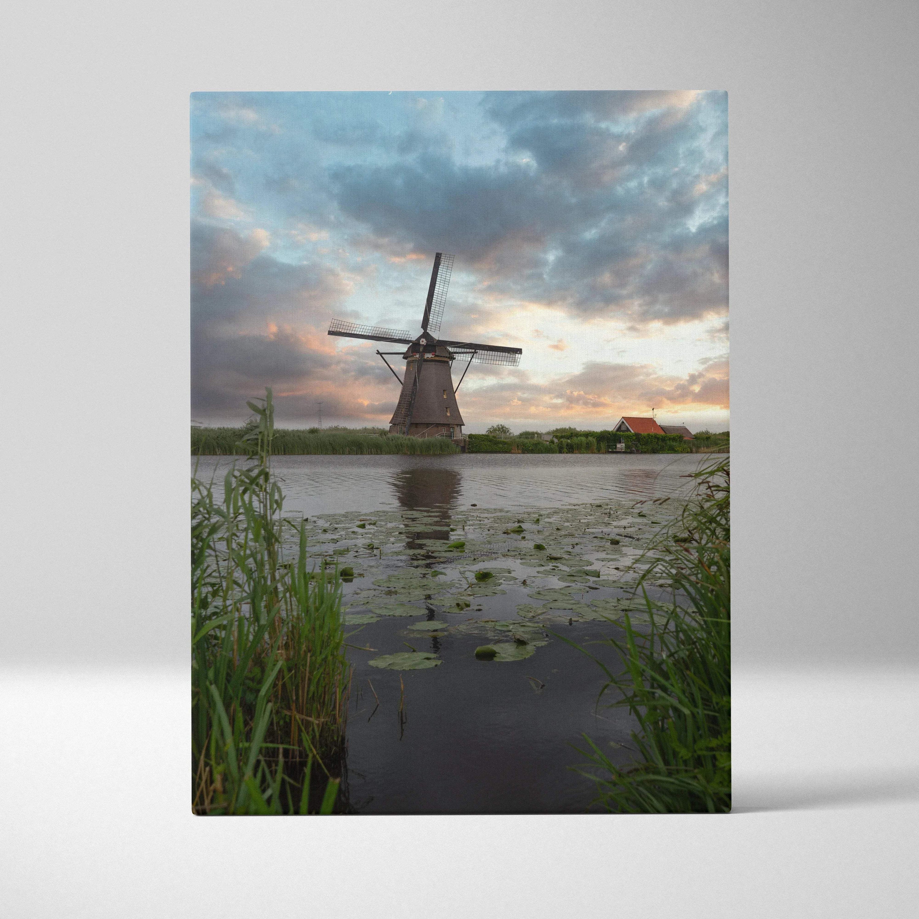 Vertical canvas wall art of a windmill at sunset over a pond with lily pads and tall grass.