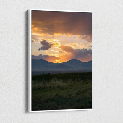 Vertical canvas wall art of a vivid sunset casting golden light over mountain silhouettes and grassy foreground.