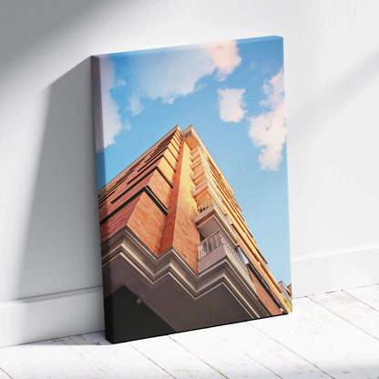 Vertical canvas wall art featuring modern urban architecture with warm brick tones against a blue sky.