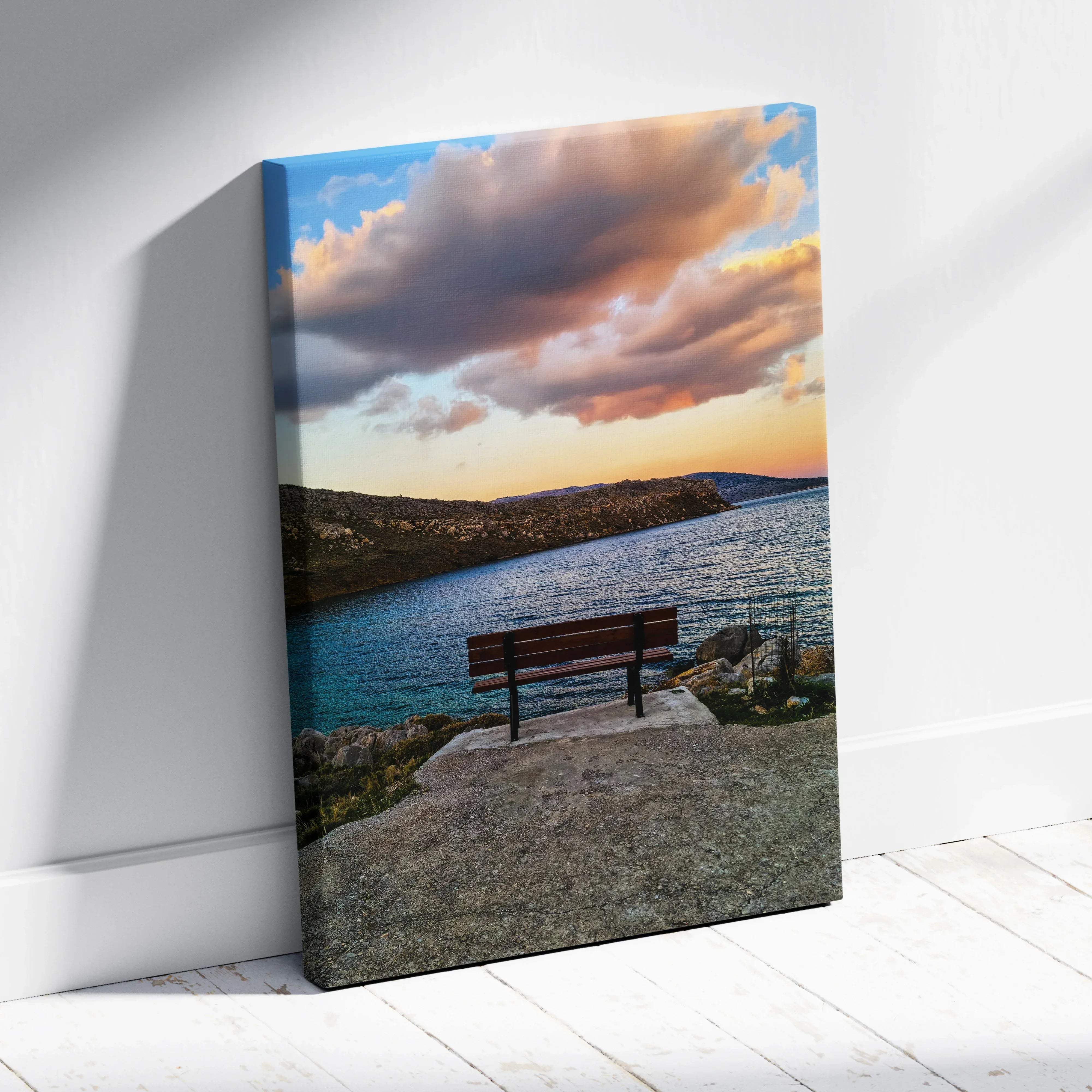 A rectangular canvas print of a wooden bench by calm blue waters under a dramatic orange and blue sunset sky