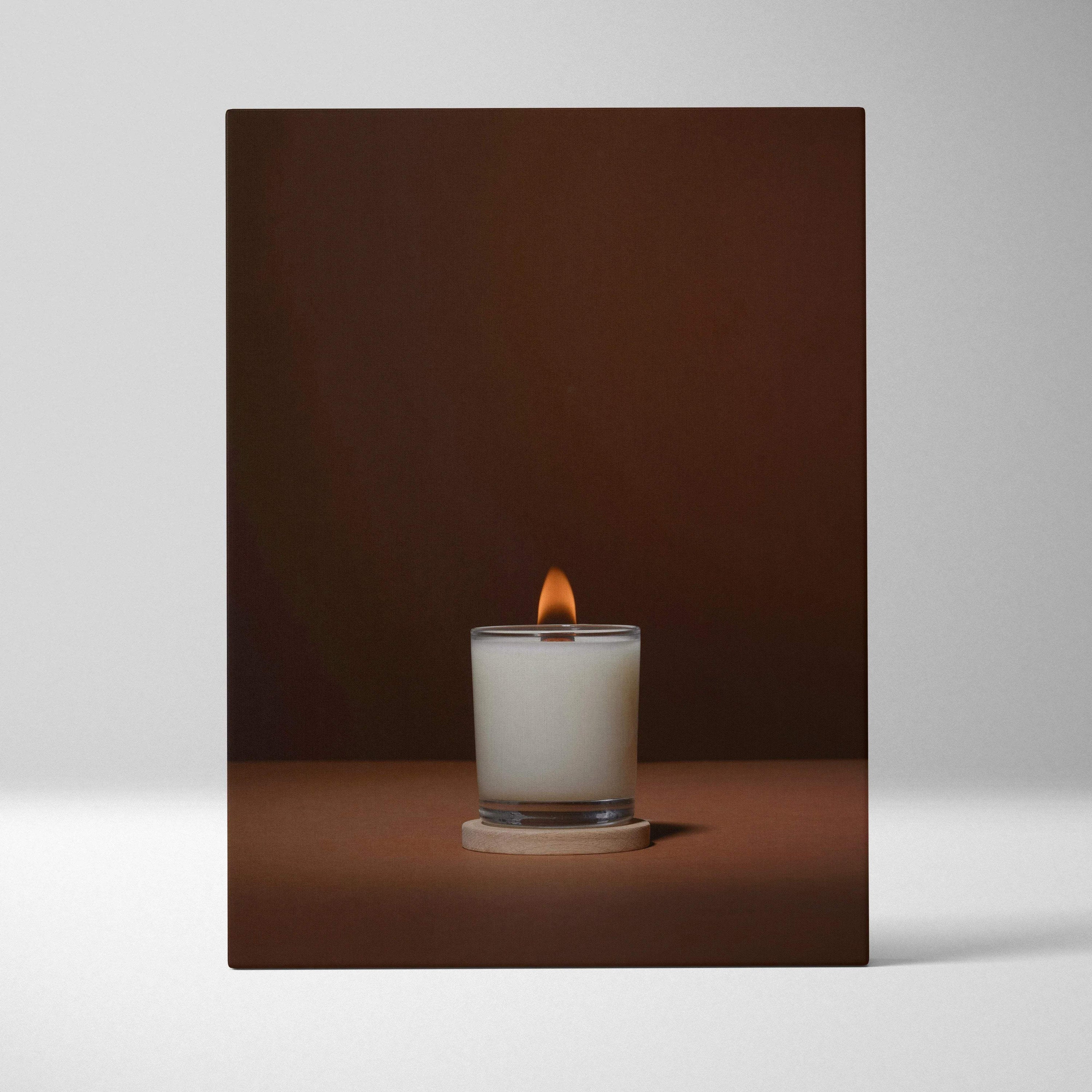 Lit white scented candle in clear glass jar on wooden coaster with warm glow