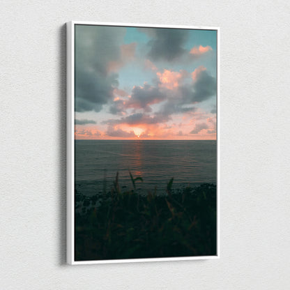 Vertical canvas wall art featuring a vibrant beach sunset over a calm ocean with dark foreground foliage.