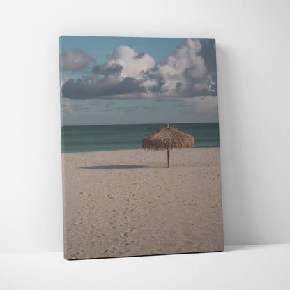 Tranquil beach scene canvas wall art with sandy shore, ocean, and thatched umbrella under cloudy sky.