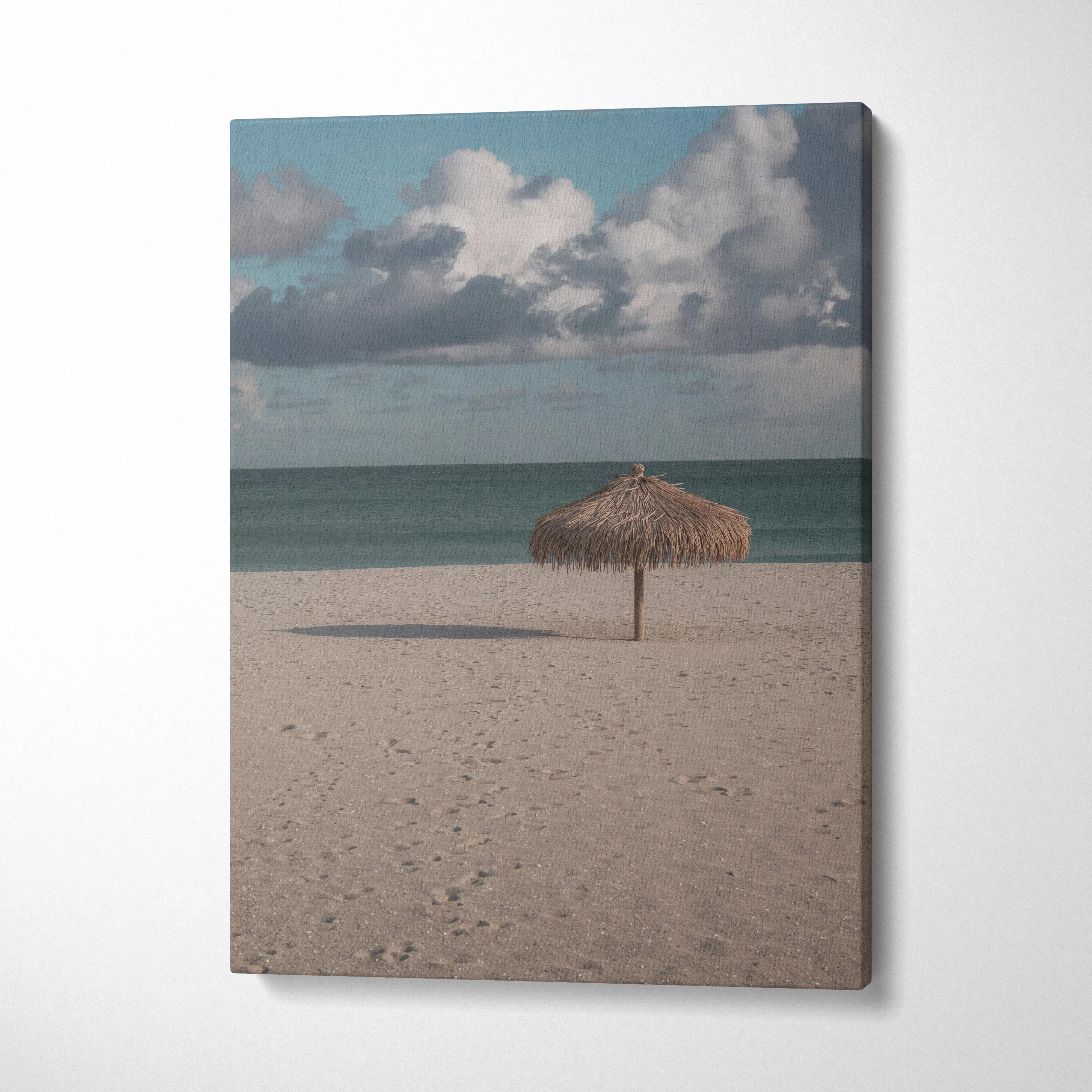 Vertical canvas wall art featuring a tranquil beach scene with a straw umbrella under a cloudy blue sky.