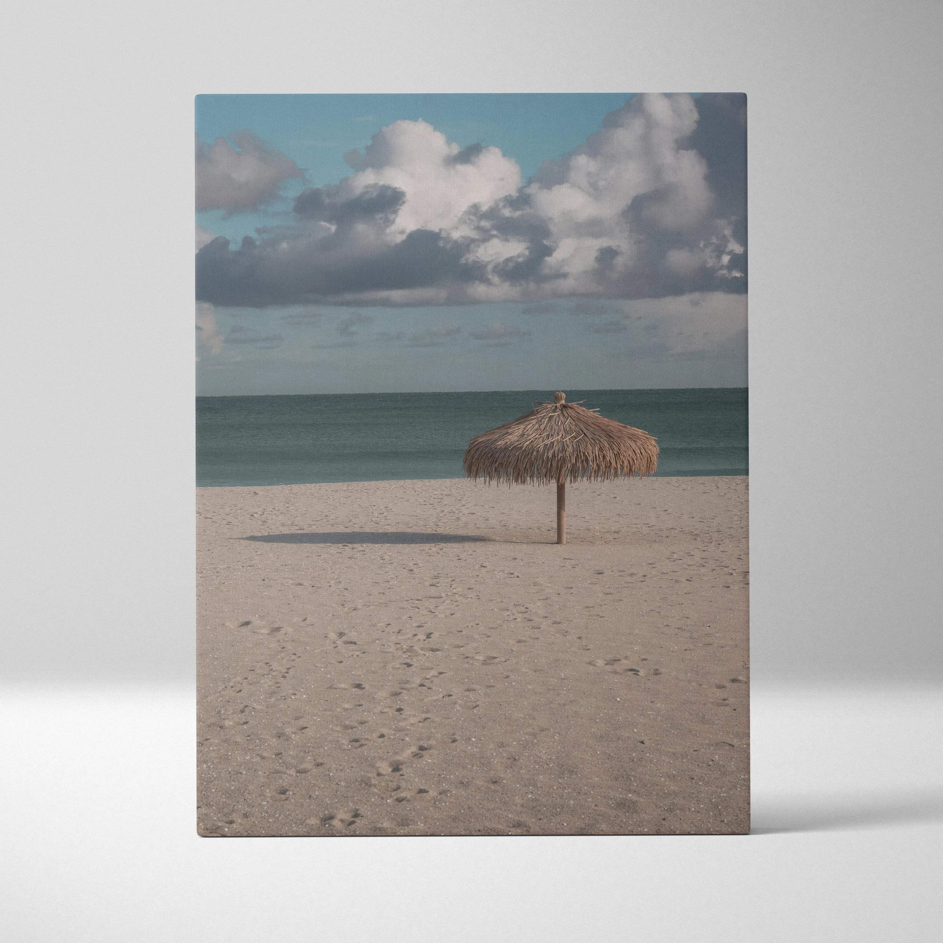 Vertical canvas wall art of tranquil beach with lone thatched umbrella under cloudy sky.
