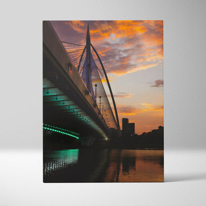 Vertical canvas wall art featuring a city skyline at sunset with illuminated bridge and reflective water.