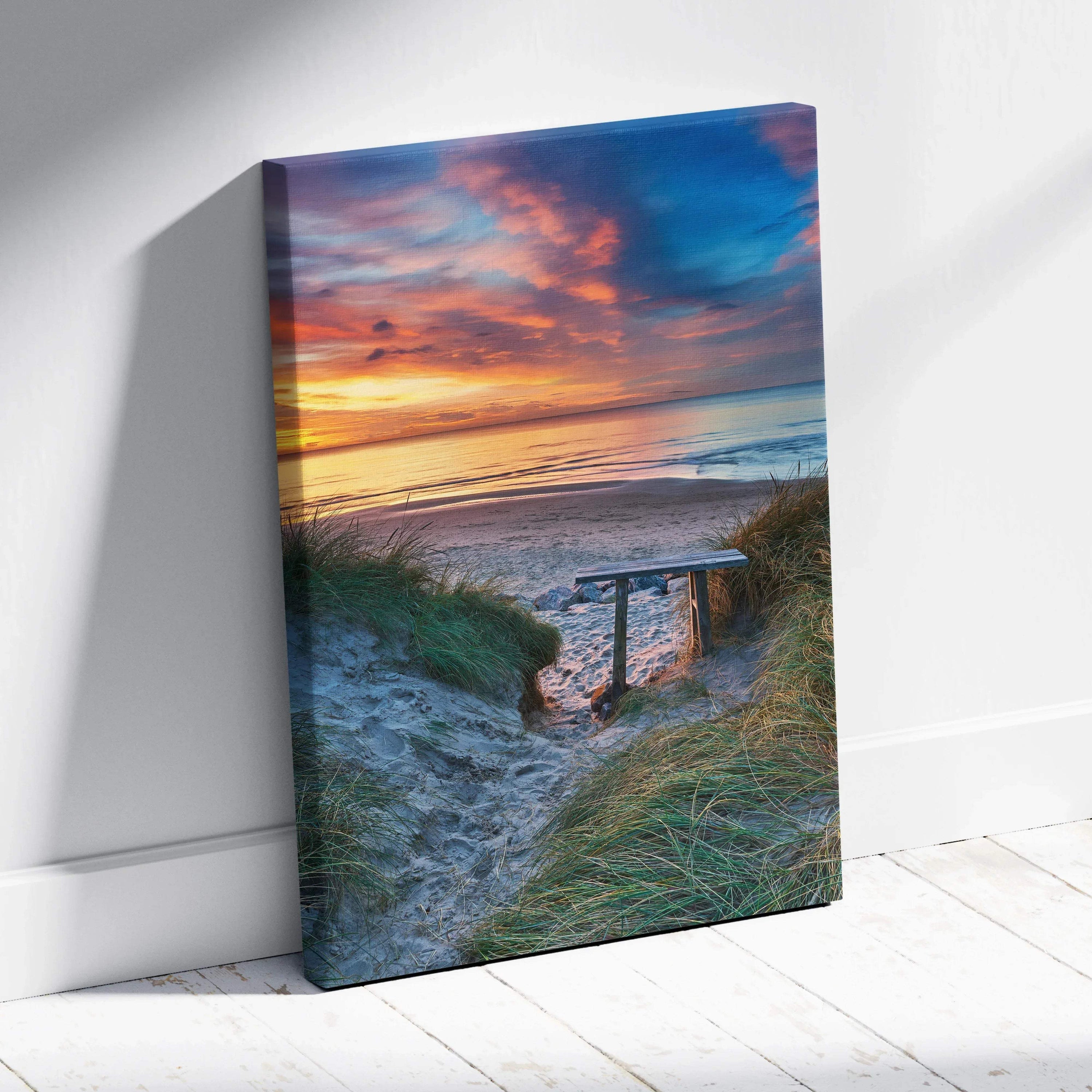 Vertical canvas wall art featuring a vibrant beach sunset with sand dunes and a wooden bench.