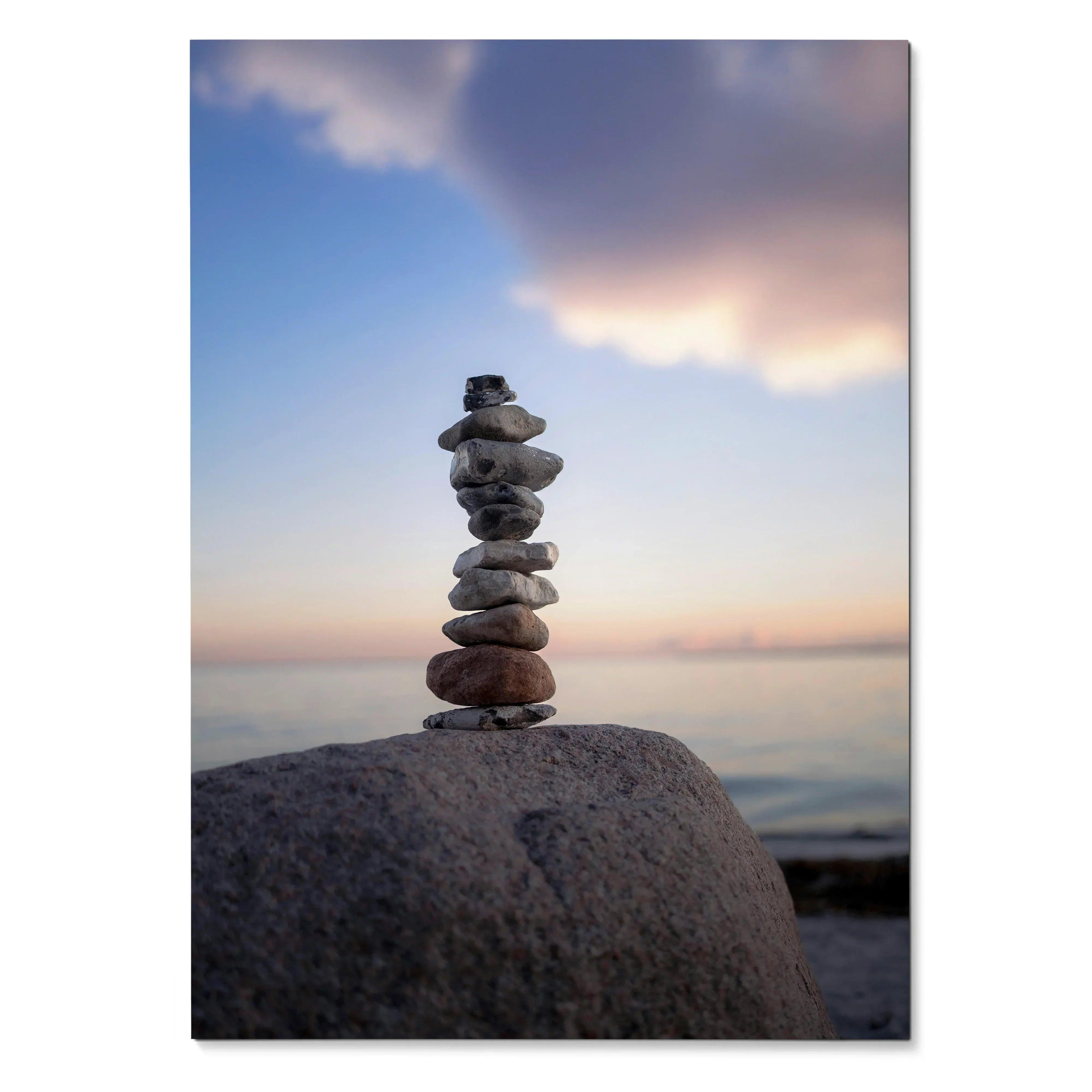 Vertical wall art poster featuring balanced stack of smooth stones on large rock by water under pastel sunset sky.