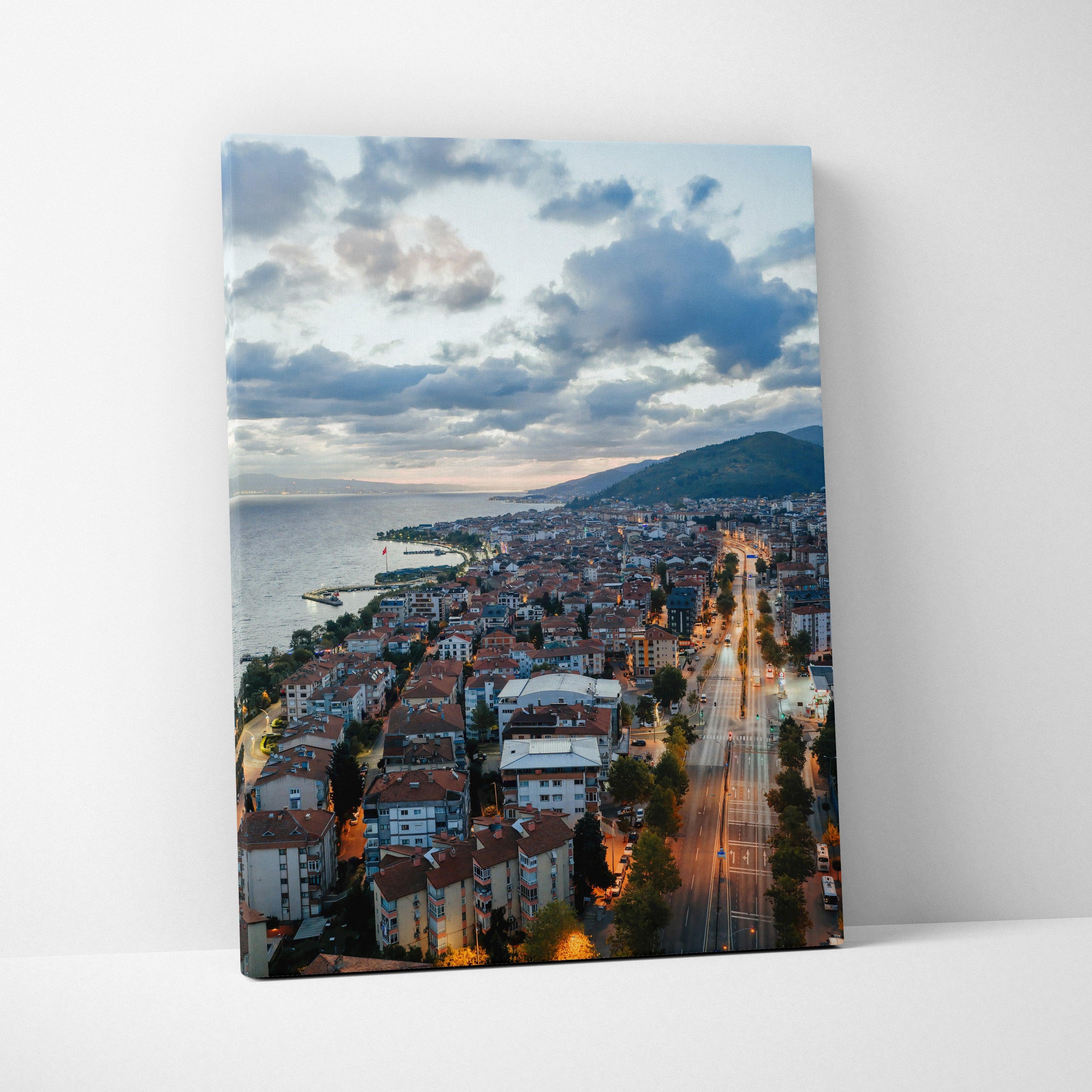 Vertical canvas wall art of a vibrant cityscape at sunset with illuminated buildings and streets under dramatic clouds.